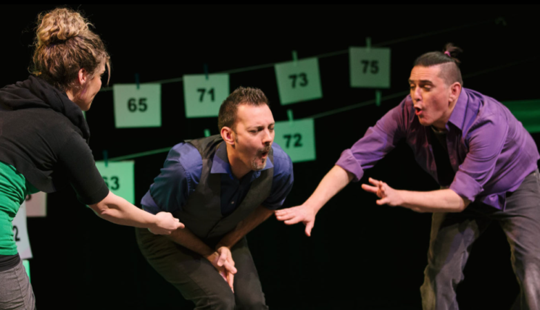 Three people perform animatedly on stage, gesturing dramatically. Numbered cards hang on strings behind them. The background is dark, highlighting the performers expressive faces and movements.